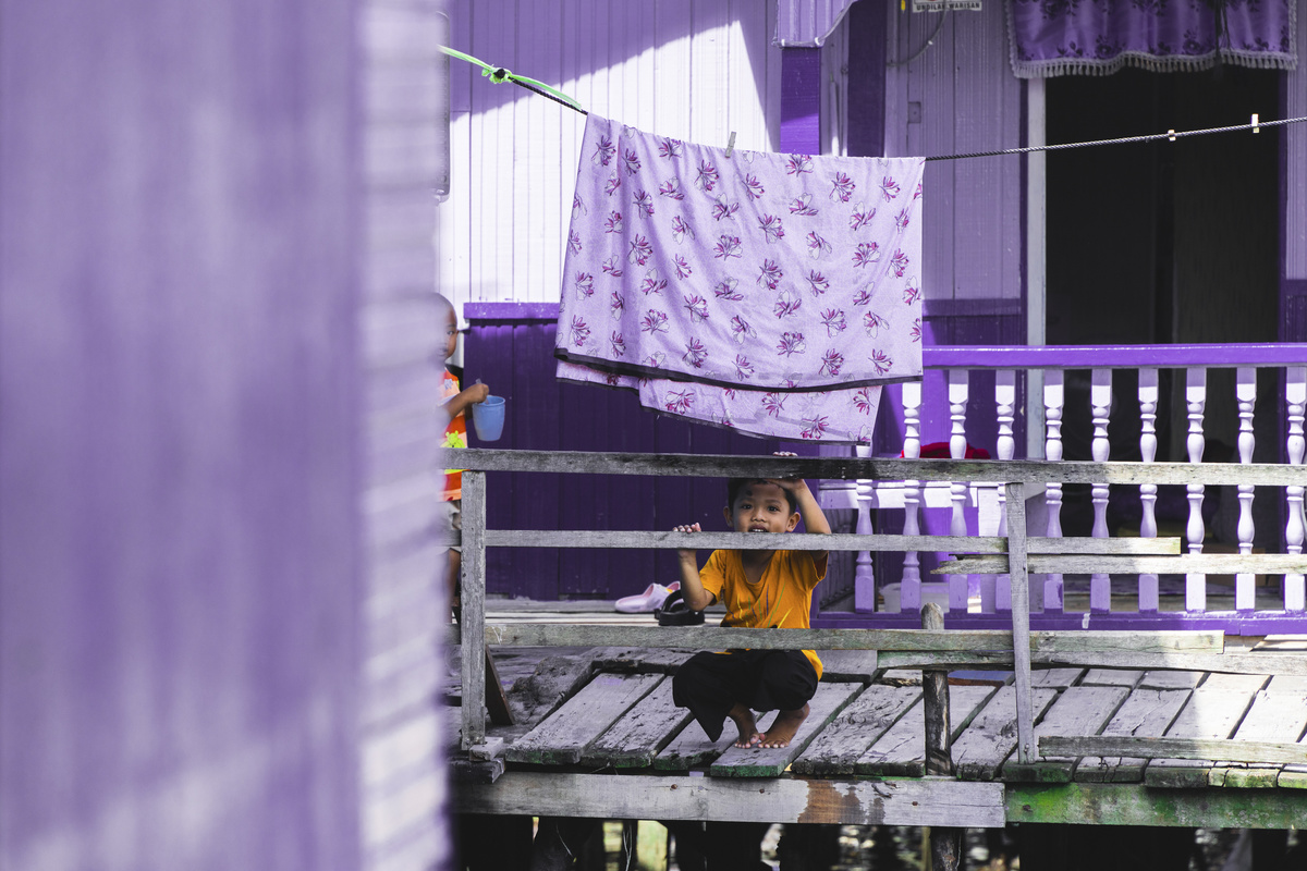 一个坐在木凳上的年轻男孩 带着一把紫色雨伞 站在一所紫色房子外面。