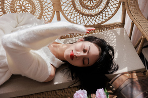 一个涂着口红的女青年躺在床上 周围有花和一把椅子。
