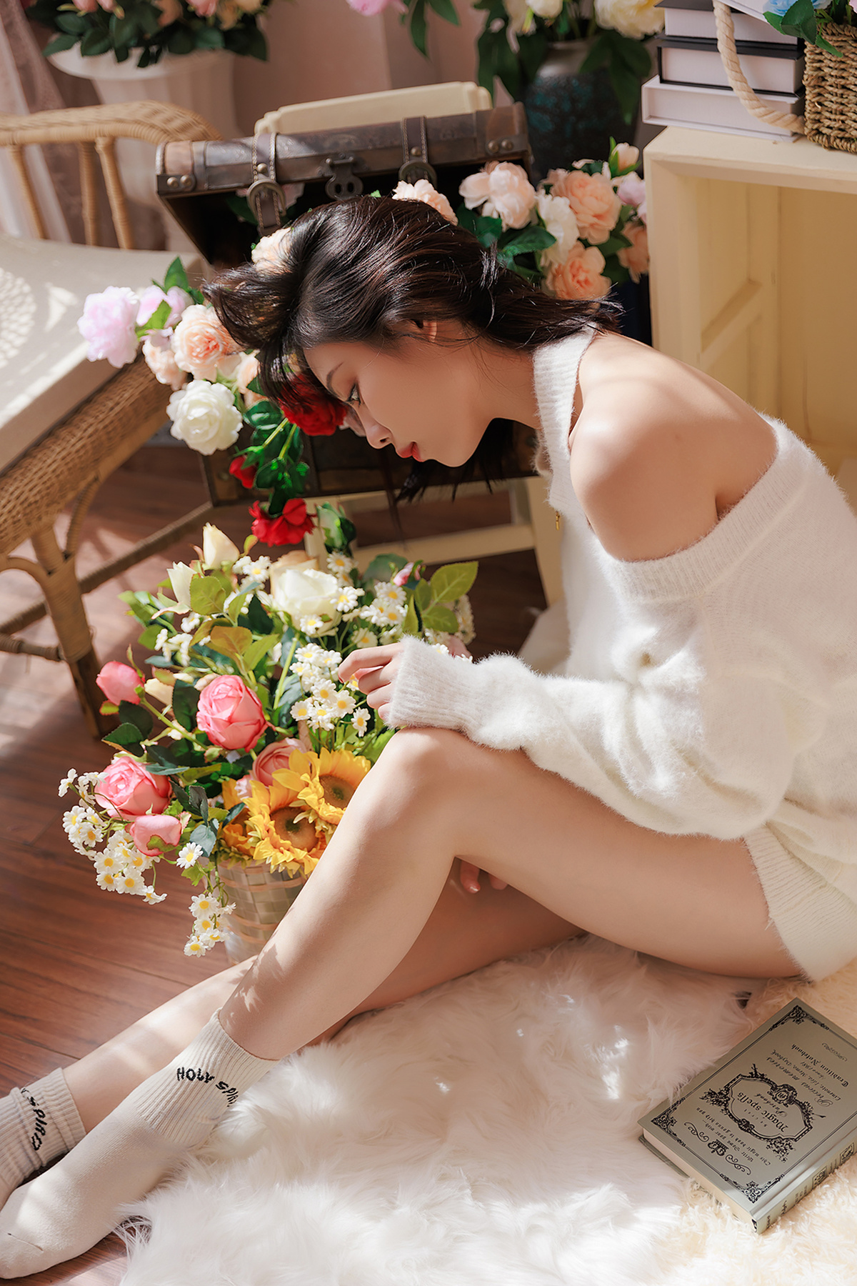 一个穿着白色连衣裙的年轻女子坐在地板上 旁边有一本书和花。