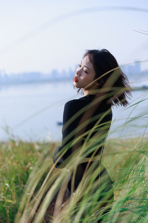 一个穿黑色衣服的年轻女子坐在一片高草丛生的草地附近的水边。