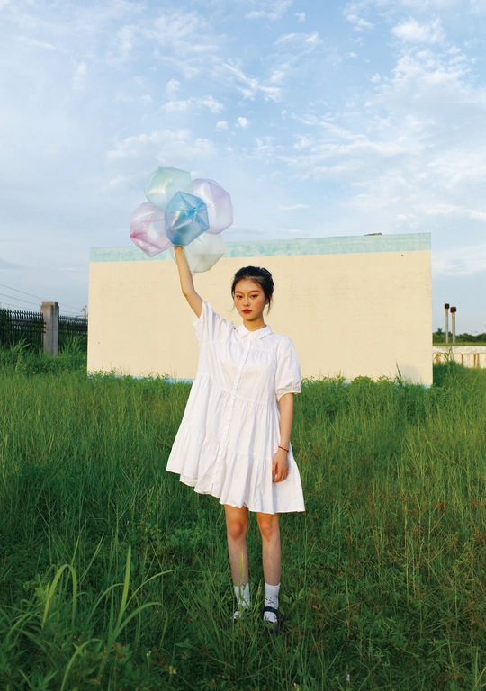 一位身穿白色连衣裙、手持蓝色气球的年轻女子站在草地上