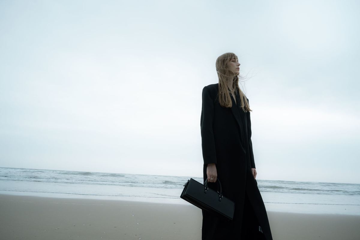 一位穿着长款外套的女性 holding a黑色手提箱在沙滩上行走