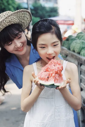 一位女子抱着一个小女孩在街上吃西瓜。