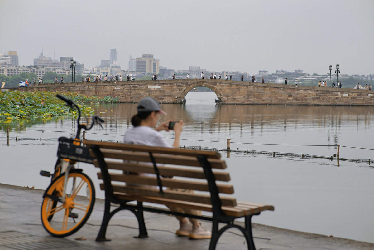一个人坐在木凳上 自行车停在上面 眺望着一处水域 那里有桥和城市在远处。