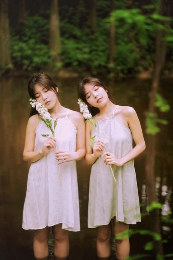 两个穿着白色连衣裙、手捧鲜花的年轻女孩在水面前摆姿势