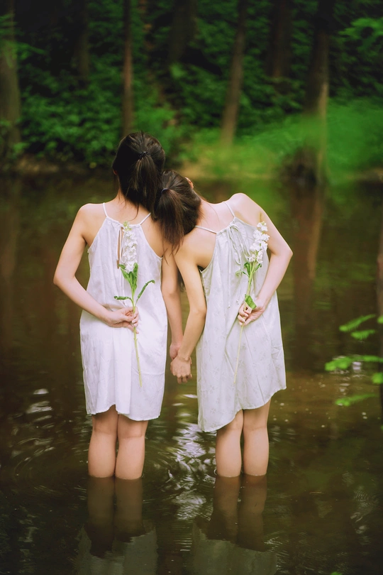 两个穿着白色连衣裙的年轻女子手牵着手穿过小溪或河流 在水中手捧鲜花