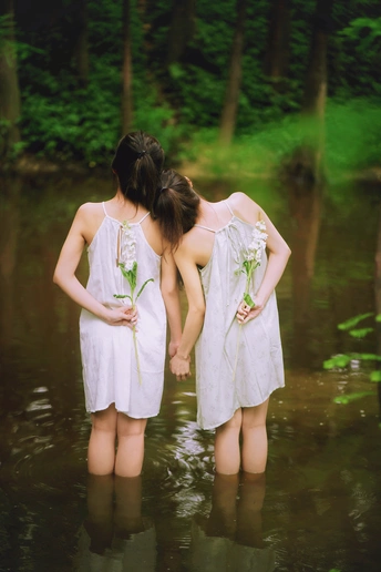 两个穿着白色连衣裙的年轻女子手牵着手穿过小溪或河流 在水中手捧鲜花