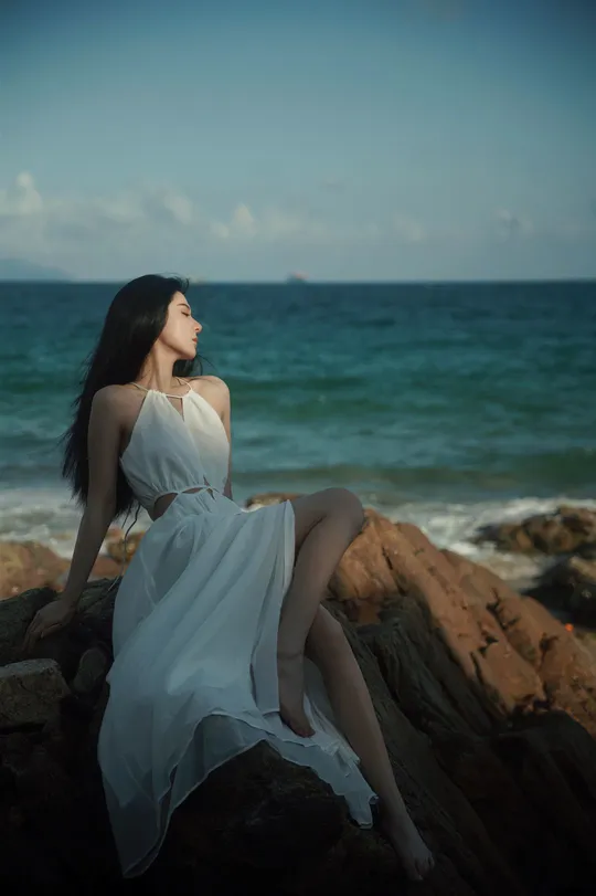 一位穿着白色连衣裙的美女坐在海边的一块岩石上