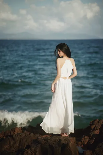 一位穿着白色连衣裙的年轻女子站在岩石海滩上 眺望着大海。
