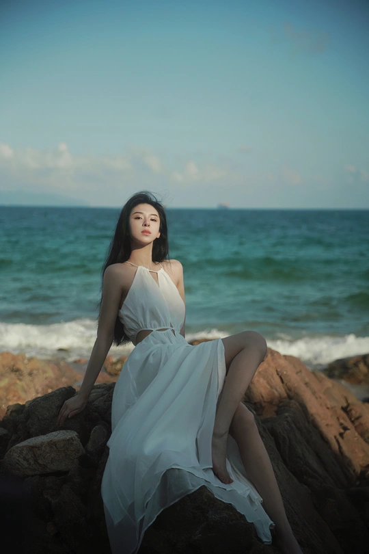 一位穿着白色连衣裙的年轻女子坐在海边或海洋边的岩石上