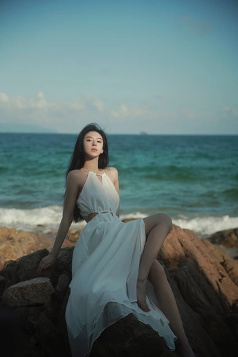 一位穿着白色连衣裙的年轻女子坐在海边或海洋边的岩石上