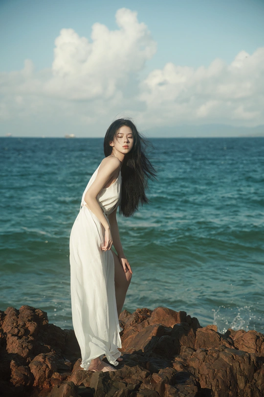 一位美丽的年轻女子穿着一件长款白色连衣裙在海洋边的一个岩石海滩上摆姿势