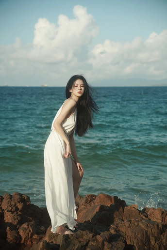 一位美丽的年轻女子穿着一件长款白色连衣裙在海洋边的一个岩石海滩上摆姿势