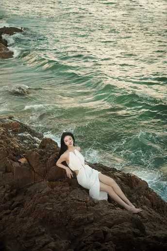 一位美丽的年轻女子穿着白色连衣裙坐在海洋边的一个岩石海滩上