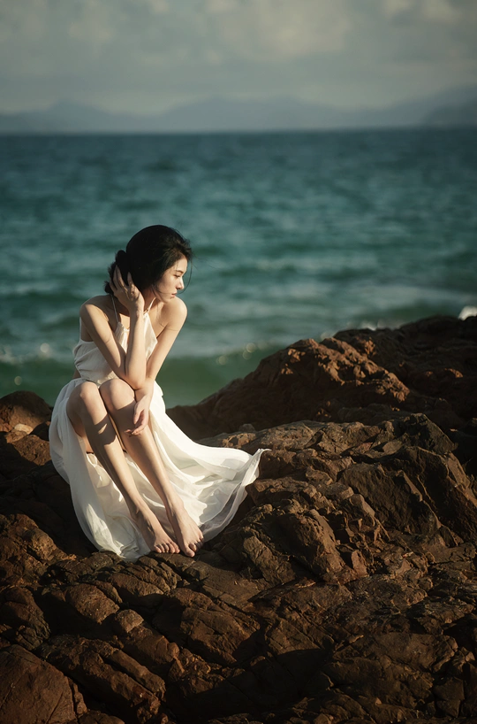 一位美丽的年轻女子穿着白色连衣裙坐在海洋边的一个岩石海滩上