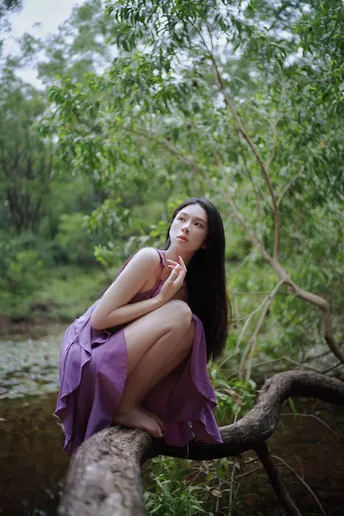 一位美丽的年轻女子穿着紫色的裙子坐在一条河流或小溪中的倒下树木的树枝上。