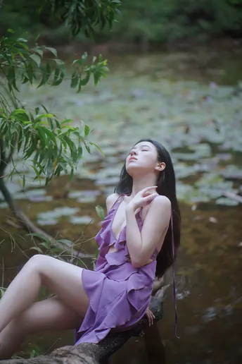 一个美丽的年轻女子穿着紫色的裙子坐在一条河流或小溪中的一棵倒下的树上