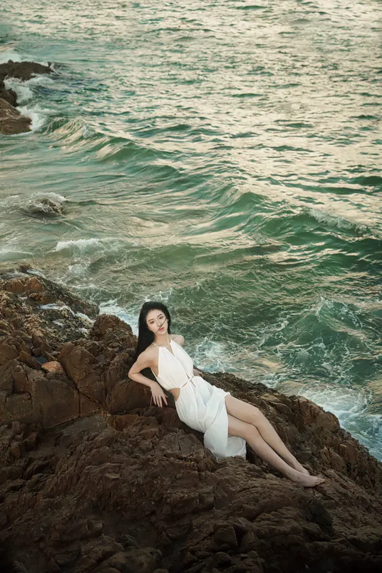 一位美丽的年轻女子穿着白色连衣裙坐在海洋边的一个岩石海滩上 海浪汹涌澎湃。