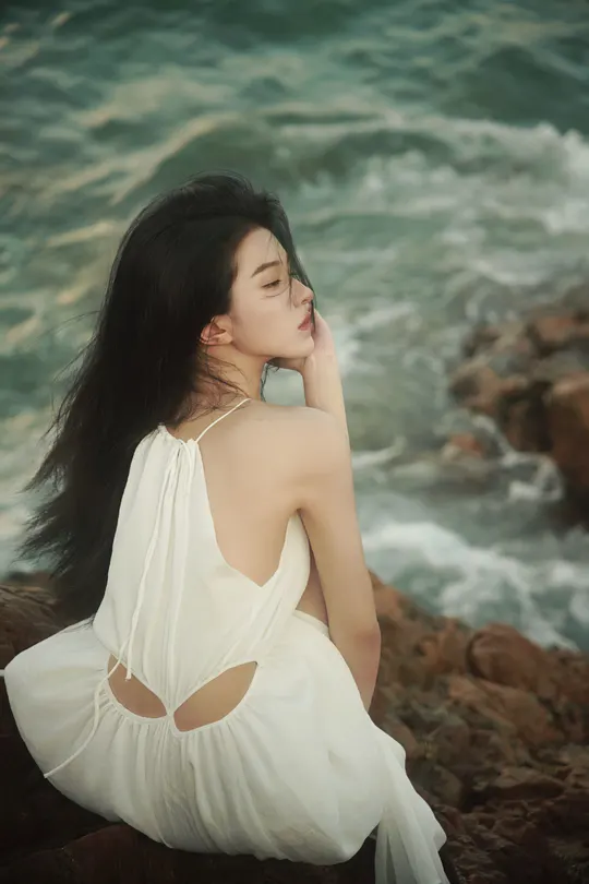 一位穿着白色连衣裙的美女坐在海边的一块岩石上