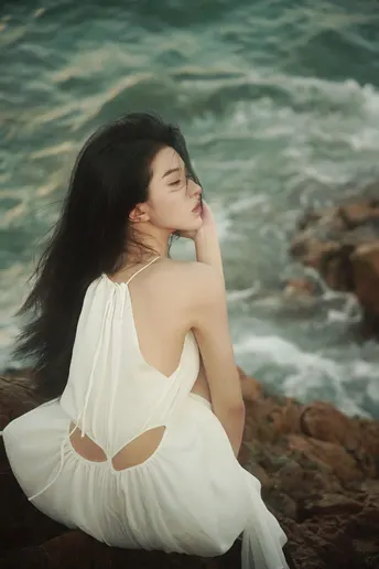 一位穿着白色连衣裙的美女坐在海边的一块岩石上