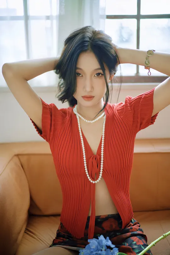 一位穿着红衬衫和项链的年轻女子坐在沙发上 旁边有一朵蓝色的花。