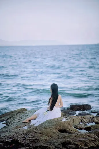 一个穿着白色连衣裙的女孩坐在岩石上 面对海洋 在岩石海滩上。