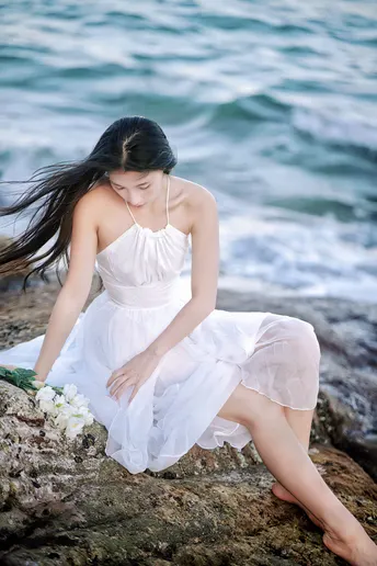 一个穿着长白色裙子的年轻女子坐在靠水边的岩石上 周围有海洋中的花朵。