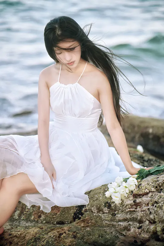 一个穿着白色连衣裙的年轻女子坐在靠近海洋的岩石上