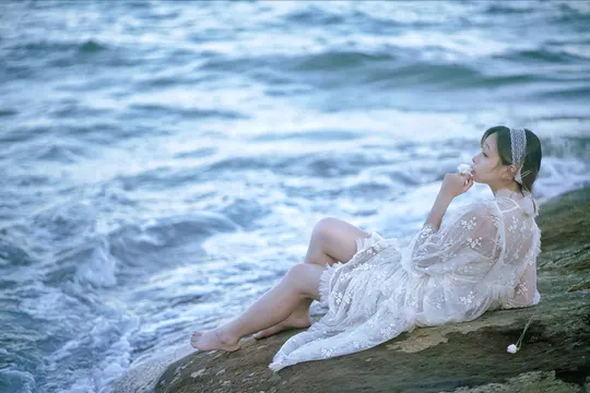 美丽的年轻女子穿着白色连衣裙坐在海滩边的一块岩石上 靠近海洋。