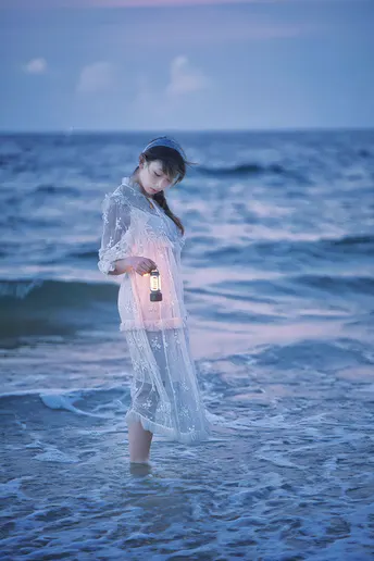 一位穿着连衣裙的年轻女子手持点燃的蜡烛站在海滩的水中