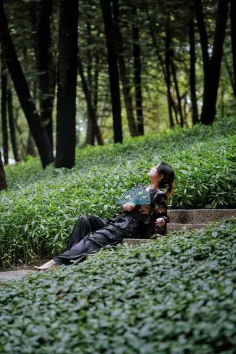 一位妇女躺在绿森林中的一张长椅上 而一位带着雨伞的男人躺在草地上。
