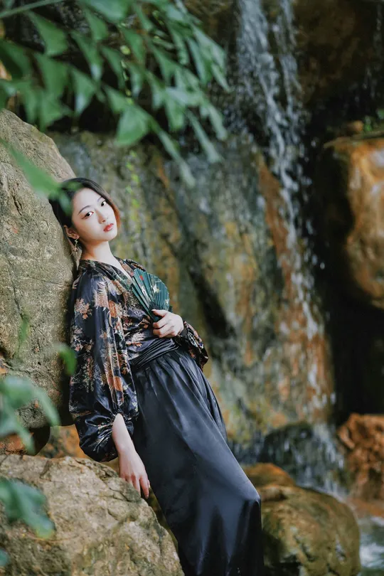 美丽的年轻女士穿着黑色连衣裙坐在靠近溪水的岩石上