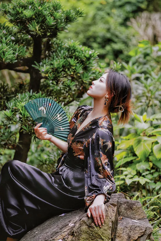 一位穿着和服的美丽年轻女子坐在岩石上 手里拿着扇子 在花园里。