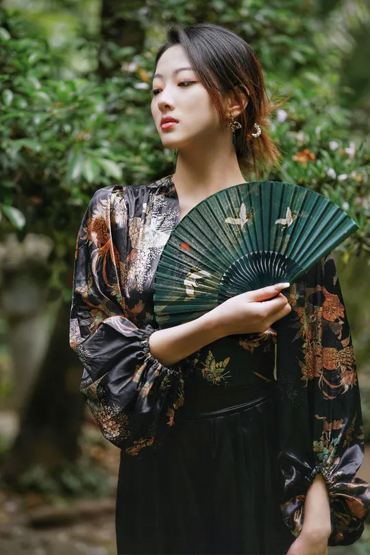 一位年轻女子穿着黑色和服手持扇子和雨伞在森林中摆姿势