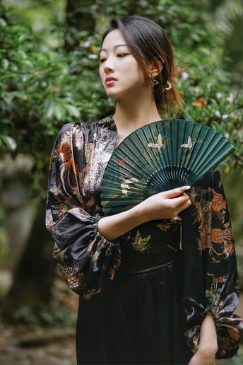 一位年轻女子穿着黑色和服手持扇子和雨伞在森林中摆姿势