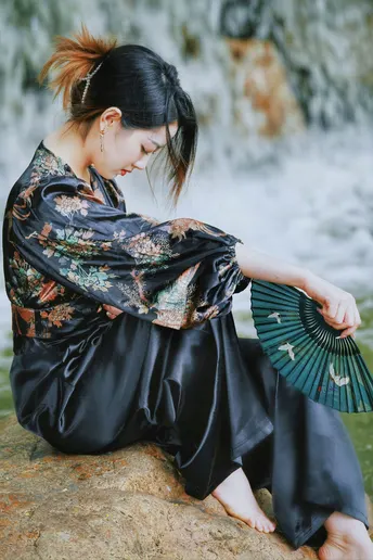 一位穿着和服的年轻女子拿着扇子坐在瀑布前的岩石上