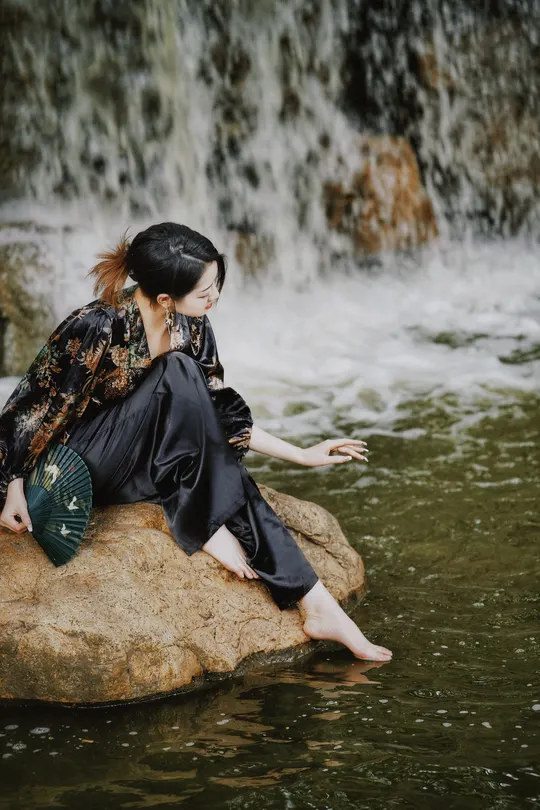 一个穿着黑色连衣裙的年轻女子坐在瀑布的水中一块岩石上