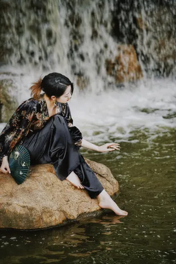 一个穿着黑色连衣裙的年轻女子坐在瀑布的水中一块岩石上