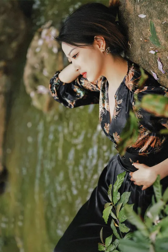 一位穿着黑色连衣裙的美女靠在森林中的一块岩石上