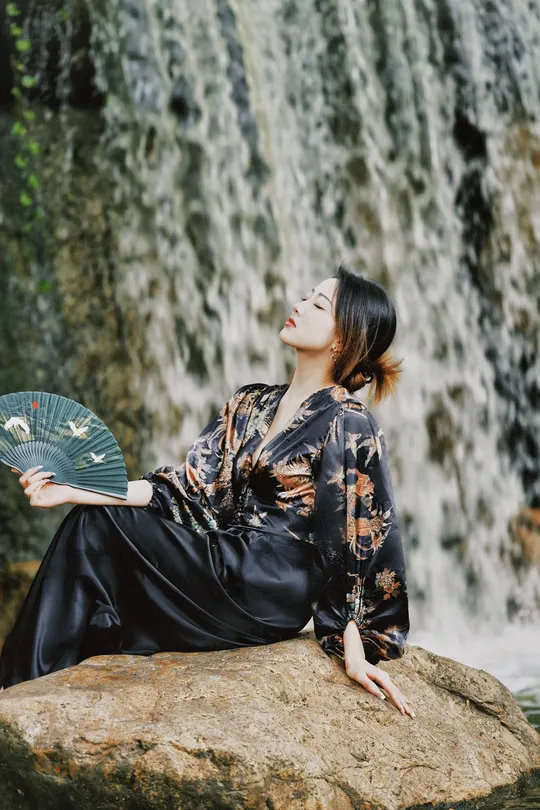 一位穿着美丽裙子的年轻女子坐在瀑布前的岩石上 手里拿着扇子。