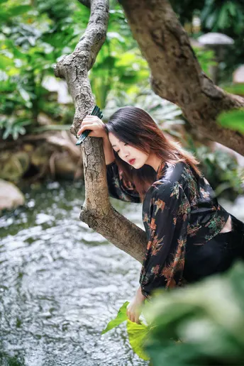 美丽年轻女子坐在树干上 旁边有一条流水