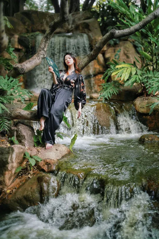 一位穿着黑色连衣裙的年轻女子 坐在瀑布旁一块岩石上 手里拿着一瓶饮料 正在摆姿势拍照。