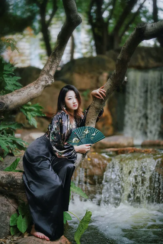 美丽的年轻女子穿着传统服装拿着扇子坐在瀑布附近的一棵树上