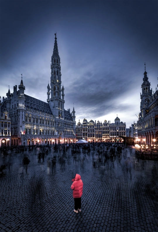 一个穿着红色外套的女人站在夜晚的广场上 周围有其他人行走。