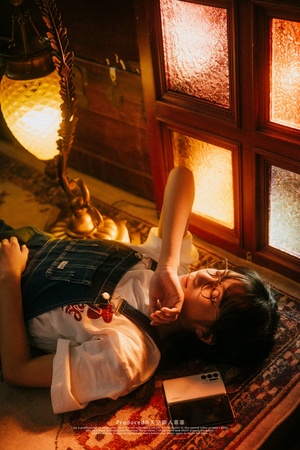 一个戴着眼镜的年轻女子躺在灯光昏暗的房间里的床上 房间里亮着灯 地板上放着一本书
