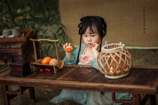 一个打扮成娃娃的小女孩坐在木桌旁 手里拿着一篮橙子和其他小水果