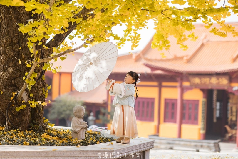 一个穿着传统服装的年轻女孩拿着一把白伞站在一棵树下