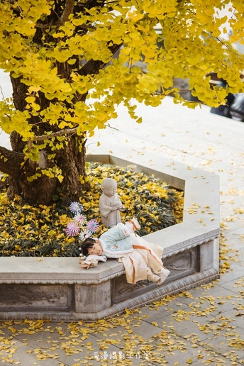 一个小女孩躺在公园里的一张长凳下 长凳下有一棵开满黄色花的树。