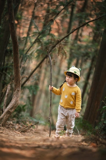 一个穿着黄色雨衣和帽子的年轻男孩拿着棍子穿过森林。