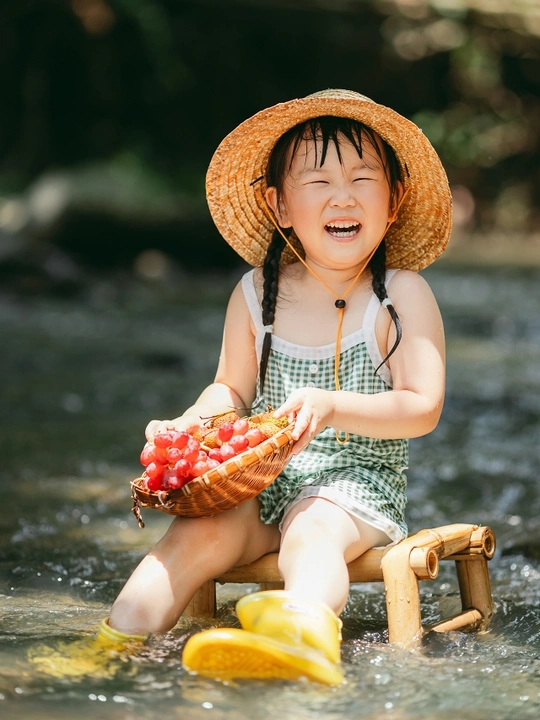 一个戴草帽的小女孩坐在木椅上 篮子里装满了葡萄 而小女孩在河里玩耍。